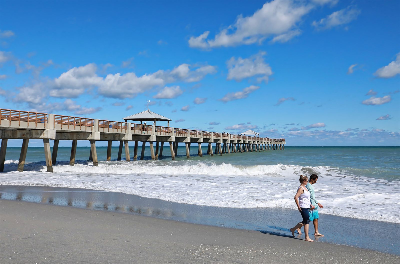 A couple walking barefoot along the surf line beside a long concrete fishing pier under a blue sky