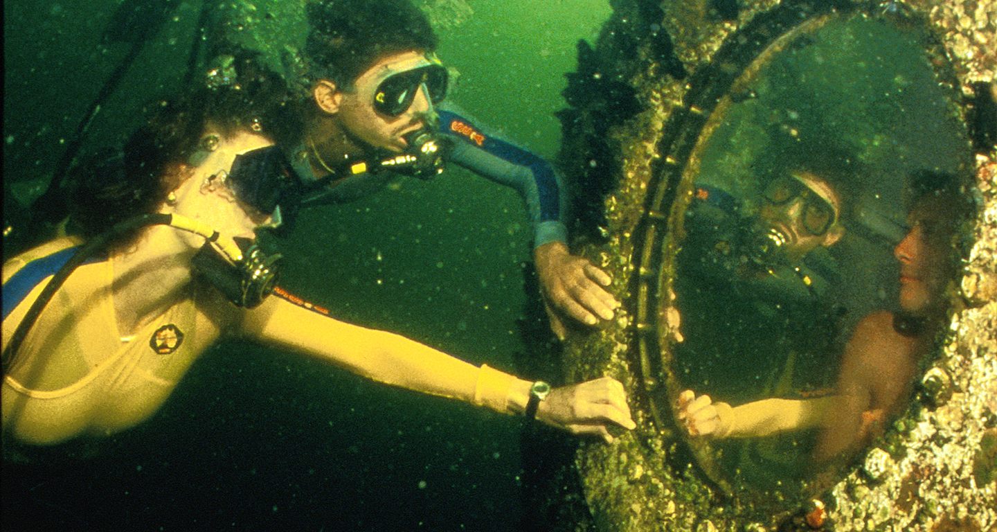 Two divers examining the porthole of a sunken shipwreck in green water